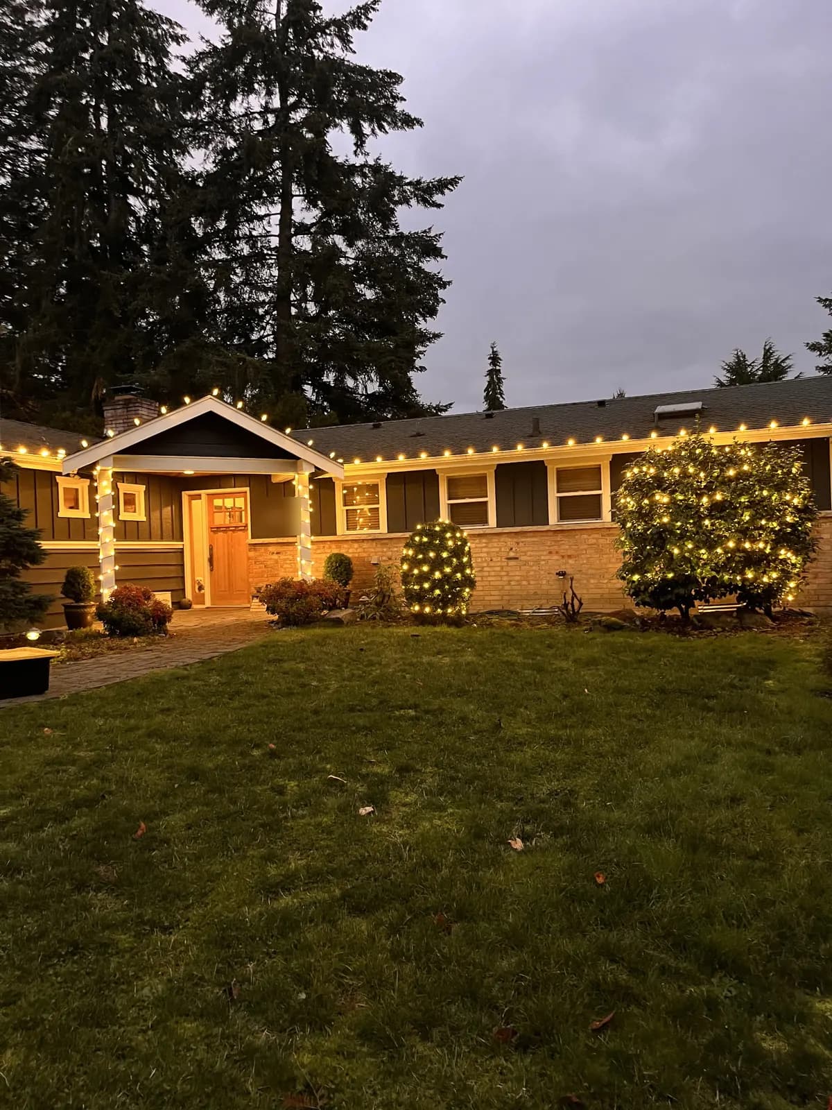Warm holiday lights running the full eave line of a craftsman home at dusk, shrubs lit below — our work